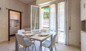 A dining area with a table set for four, a kitchen with cabinets, and a sliding glass door leading to an outdoor area. - Hostdomus - Gassa d'amante (Finale Ligure)