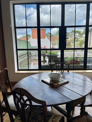 A round wooden table with a bowl and a book, a chair, and a window with a view of a garden and buildings. - Apartment Sleeps 6 With Parking (Budleigh Salterton)