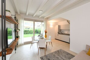 Ein modernes Wohnzimmer mit einem Glastisch, weißen Stühlen, einer Couch, einem Fernseher und einer Küchenzeile mit weißen Schränken.