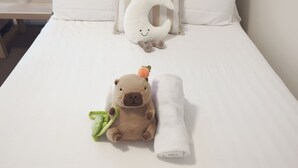 Un letto con lenzuolo bianco, un cuscino a forma di luna, un peluche a forma di orso e un asciugamano bianco arrotolato.