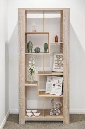 En bokhylla i trä med flera hyllplan, varav några innehåller dekorativa föremål som en kaktus, en vas med blommor och en nallebjörn.