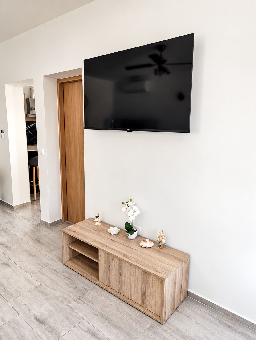 Un salon moderne avec un téléviseur à écran plat fixé au mur, une console en bois agrémentée d’objets décoratifs et un sol carrelé de couleur claire.