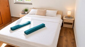 Ein ordentlich bezogenes Bett mit weißen Leinentüchern und zwei aufgerollten Handtüchern, eine Nachttischlampe, ein kleiner Topf mit Pflanze und ein Holzschrank.