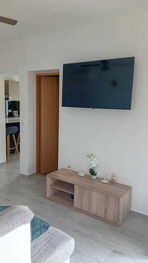 Ein modernes Wohnzimmer mit Flachbildfernseher an der Wand, einem Holzkonsolentisch mit Dekoration und einer Sitzecke mit einem gemusterten Hocker.
