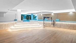 Un espace piscine intérieur moderne, avec un escalier en marbre menant à la piscine, un coin salon avec des fauteuils bleus et une fontaine décorative fixée au plafond.