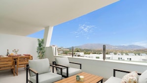 Un balcon avec une table en bois, deux chaises, une petite plante et une vue sur le paysage urbain et les montagnes.
