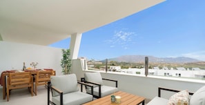 A balcony with a wooden table, two chairs, a small plant, and a view of a cityscape and mountains. - Casa Banzu | Pool - Terrace - Gym (Mijas)