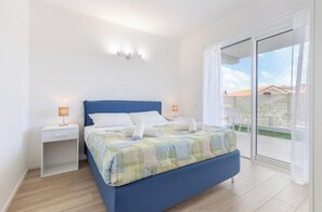 Un dormitorio con una cama azul, mesitas de noche blancas y vistas a través de un ventanal corredero.