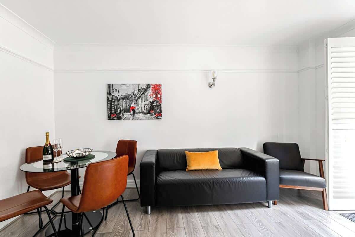 Un salon moderne avec un canapé en cuir noir, une table ronde en verre entourée de chaises, un tableau accroché au mur et une porte avec un store blanc.