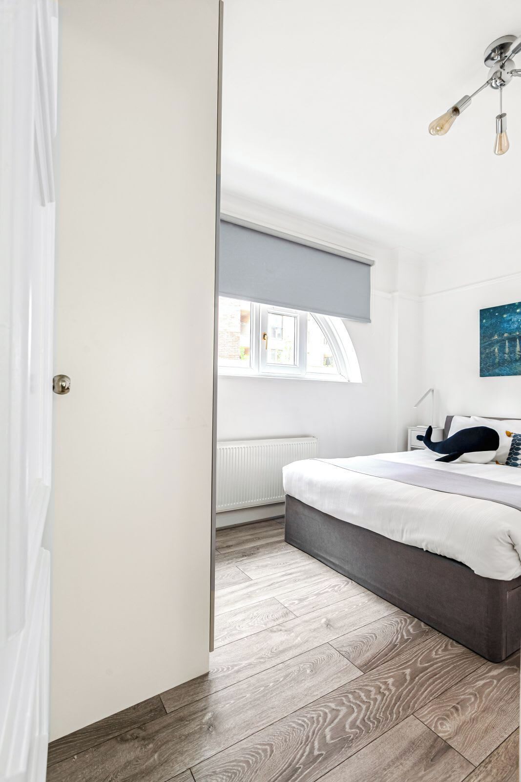 A bedroom with a bed, a radiator, a window with a roller blind, and a painting on the wall.