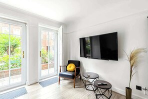 Ein Zimmer mit an der Wand montiertem Fernseher, einem Sessel mit Kissen, einem kleinen Tisch und Blick durch eine Glastür.
