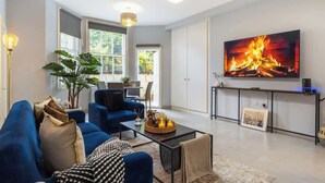 A modern living room with a large TV, blue sofas, a coffee table, and a potted plant. - 1-Bed & Patio - 2 Min Walk to Abbey Road Studios (Greater London)