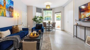 A modern living room with a blue sofa, a dining area with a table and chairs, and a large window with a view of greenery. - 1-Bed & Patio - 2 Min Walk to Abbey Road Studios (Greater London)
