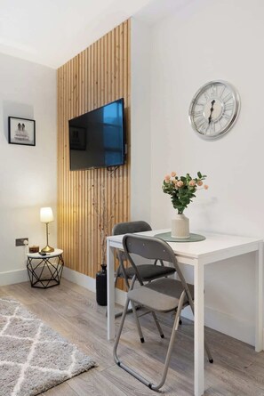 Ein modernes Zimmer mit einer Wand in Holzoptik, einem Flachbildfernseher, einer Wanduhr, einem Esstisch mit Stühlen, einer Lampe und einer Vase mit Blumen.