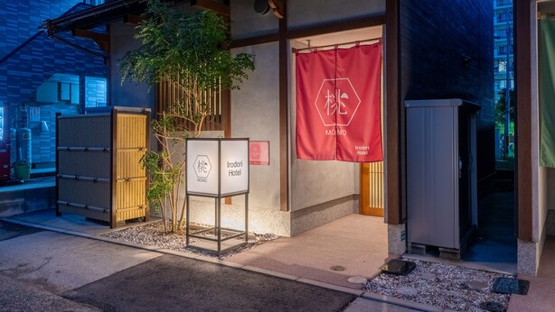 A traditional Japanese building with a red banner, a signboard, and a small tree. - Irodori Hotel MOMO (fukuokashi,hakataku,katakasu)