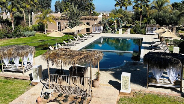 A pool area with lounge chairs, a thatched-roof structure, and a view of the ocean. - Les Villas du Douar (Marrakech)