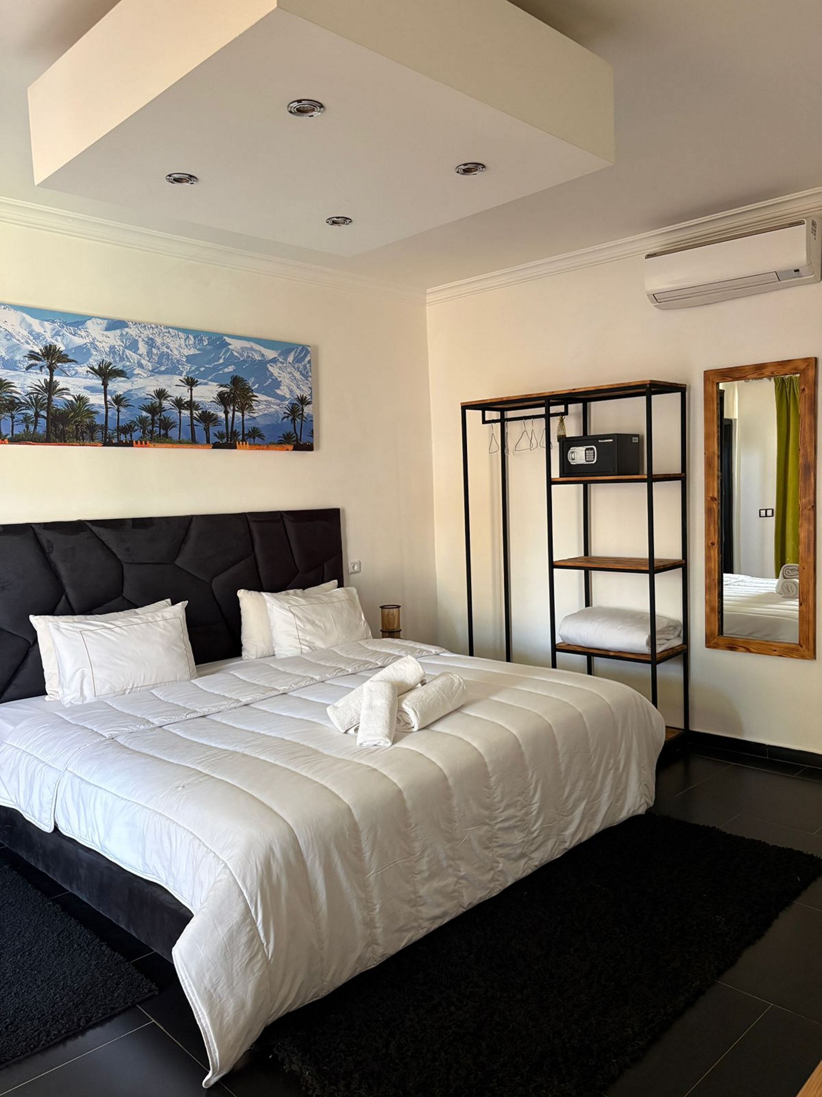 Ein Hotelzimmer mit einem großen Bett, einem Gemälde von einer Berglandschaft, einem schwarzen Metallregal und einem Spiegel mit grünem Vorhang.