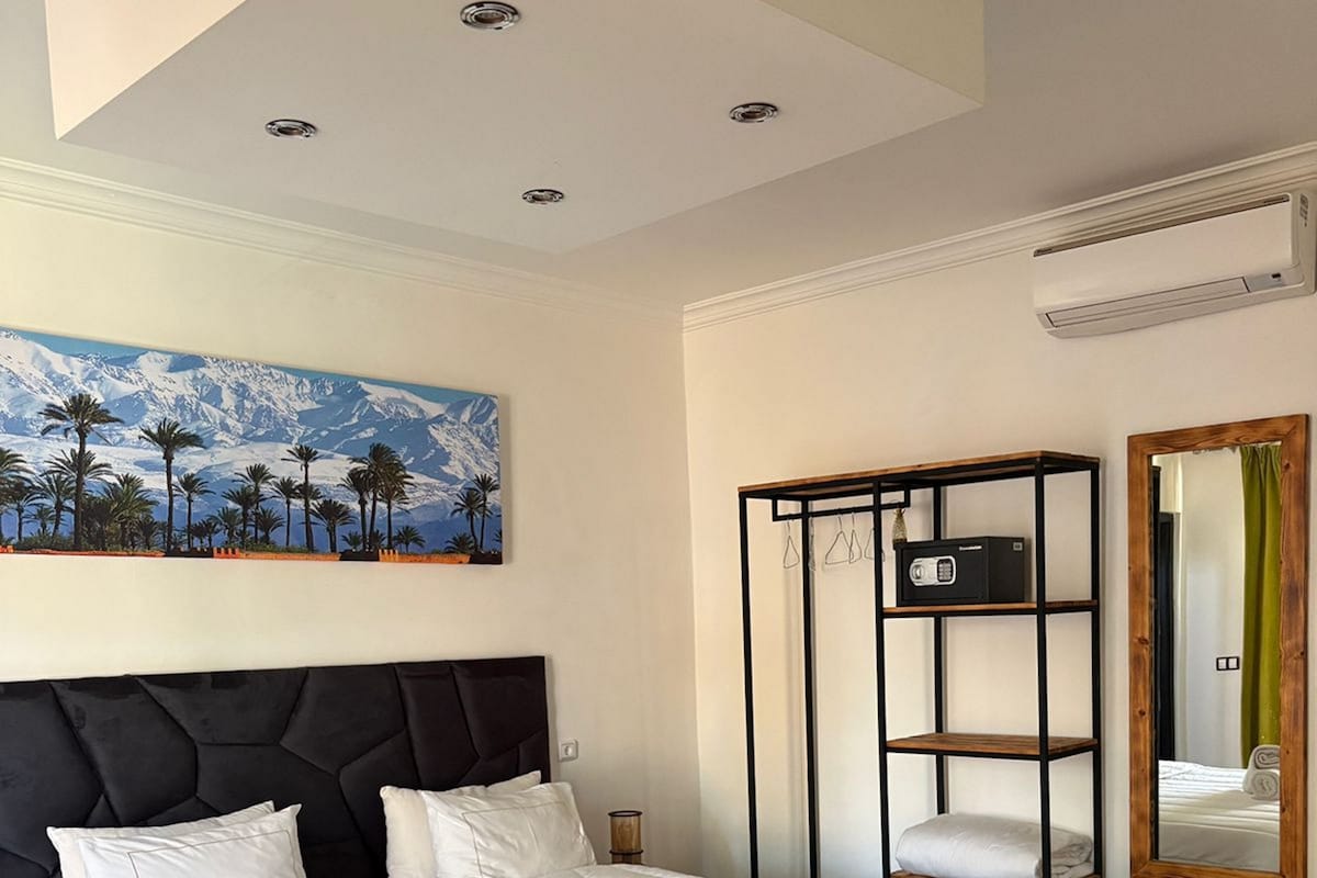 Una habitación de hotel con una cama grande, un cuadro de un paisaje montañoso, un estante de metal negro y un espejo con cortina verde.