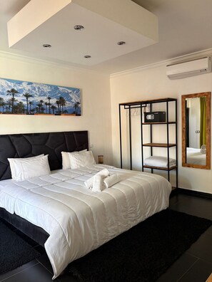 Ein Hotelzimmer mit einem großen Bett, einem Gemälde von einer Berglandschaft, einem schwarzen Metallregal und einem Spiegel mit grünem Vorhang.