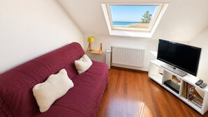 A room with a red sofa, a television on a white stand, a radiator, and a window with a view of the sea. - Duplex sea view, pets welcome, parking - 6 guests (Merville-Franceville-Plage)