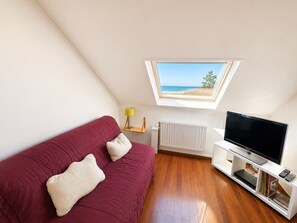 A room with a red sofa, a television on a white stand, a radiator, and a window with a view of the sea. - Duplex sea view, pets welcome, parking - 6 guests (Merville-Franceville-Plage)