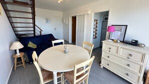 A dining area with a round table, chairs, a lamp, a sofa, a dresser, and a ceiling light fixture. - Duplex sea view, pets welcome, parking - 6 guests (Merville-Franceville-Plage)