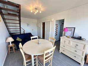 A dining area with a round table, chairs, a lamp, a sofa, a dresser, and a ceiling light fixture. - Duplex sea view, pets welcome, parking - 6 guests (Merville-Franceville-Plage)