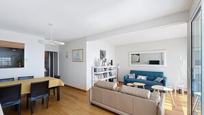 Ein modernes Wohnzimmer mit einer Couch, einem Esstisch, Stühlen, einem Bücherregal und einem großen Fenster.