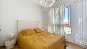 Ein Schlafzimmer mit einem Bett, einem Nachttisch, einem Sessel, einem Fenster mit Vorhängen und einer Deckenleuchte.