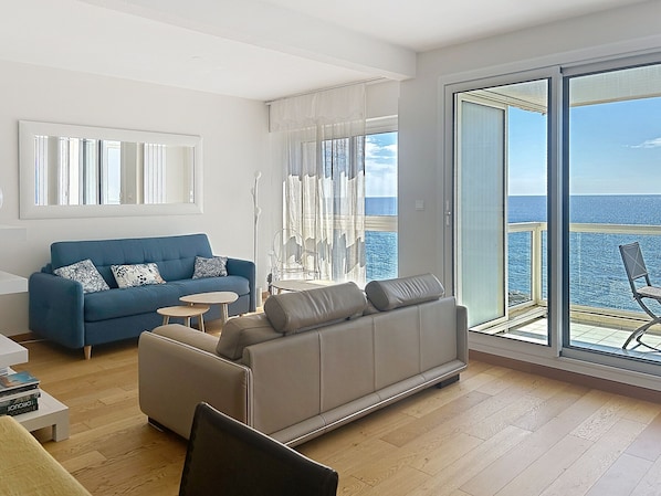 Un salon moderne avec un canapé bleu, un canapé gris et une petite table ronde. Une grande porte-fenêtre coulissante donne accès à un balcon avec vue sur la mer.