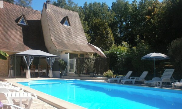 Ein Schwimmbecken mit Liegestühlen und Sonnenschirmen, eine überdachte Terrasse und ein Haus mit Satteldach.