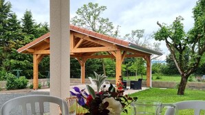 Ein überdachter Außenbereich mit Tisch und Stühlen, einer Holz-Pergola und Blick auf einen Park mit Bäumen und einem Gebäude im Hintergrund.