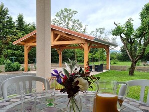 Ein überdachter Außenbereich mit Tisch und Stühlen, einer Holz-Pergola und Blick auf einen Park mit Bäumen und einem Gebäude im Hintergrund.