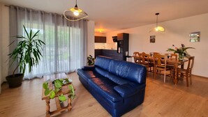 Ein Wohnzimmer mit blauem Sofa, einem Holzespeisetisch und Stühlen, einer Küchenzeile und einem großen Fenster mit durchsichtigen Vorhängen.