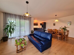 Ein Wohnzimmer mit blauem Sofa, einem Holzespeisetisch und Stühlen, einer Küchenzeile und einem großen Fenster mit durchsichtigen Vorhängen.