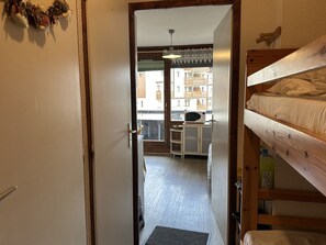Ein schmaler Flur führt zu einem Raum mit einem Etagenbett, einer Mikrowelle auf einem kleinen Tisch und Blick auf Gebäude im Freien.