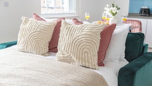 A bed with a beige and pink pillow arrangement, a teal upholstered headboard, and a window with a view of buildings. - Bright 1BR Near Piccadilly Circus (London)