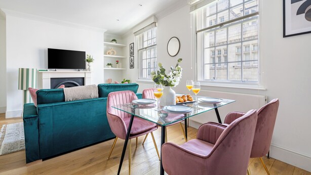 A modern living room with a teal sofa, pink chairs, a glass coffee table, and a flat-screen TV. - Bright 1BR Near Piccadilly Circus (London)