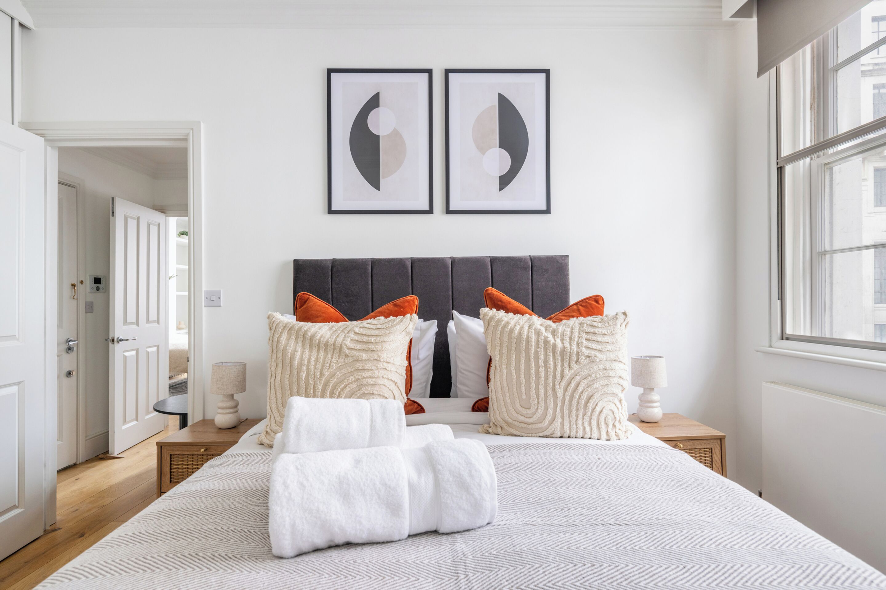 Un dormitorio con cama, almohadas, una mesita de noche, una lámpara y dos cuadros en la pared.