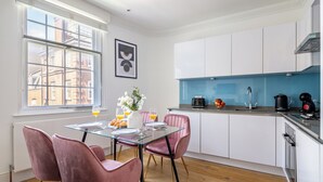 A modern kitchen with white cabinets, a glass dining table, and pink chairs. - Bright 1BR Near Piccadilly Circus (London)
