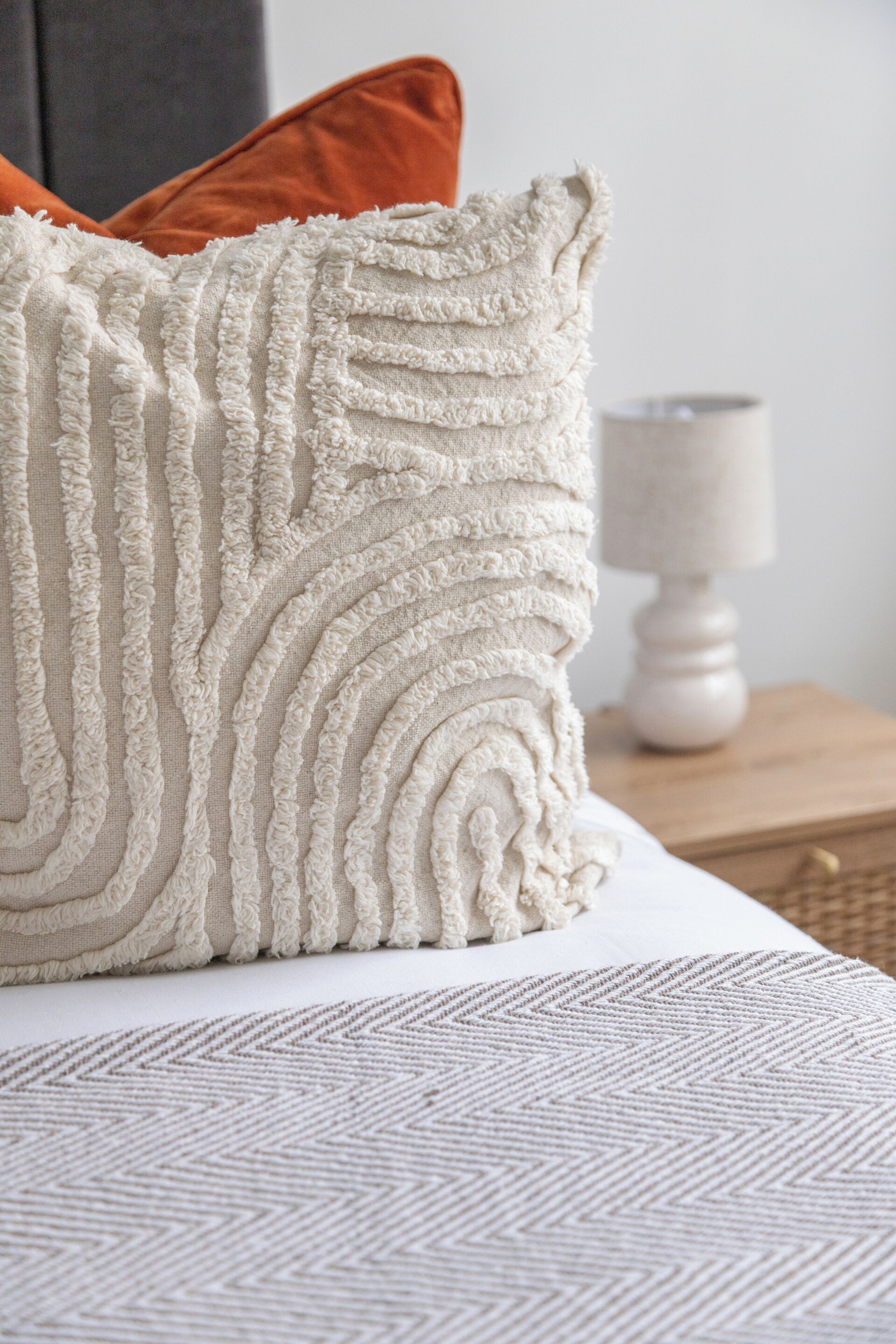 A bed with a textured white pillow, a geometric patterned blanket, and a wooden nightstand with a lamp.