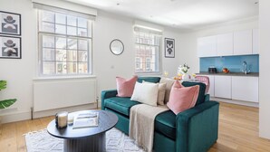 A modern living room with a teal sofa, a coffee table, and a kitchen area with white cabinets and a blue countertop. - Bright 1BR Near Piccadilly Circus (London)