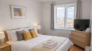 Un dormitorio con cama, mesitas de noche, televisor, ventana y vista a la ciudad.