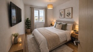 Un dormitorio con cama, mesitas de noche, televisión, ventana con cortinas, una planta y una obra de arte en la pared.