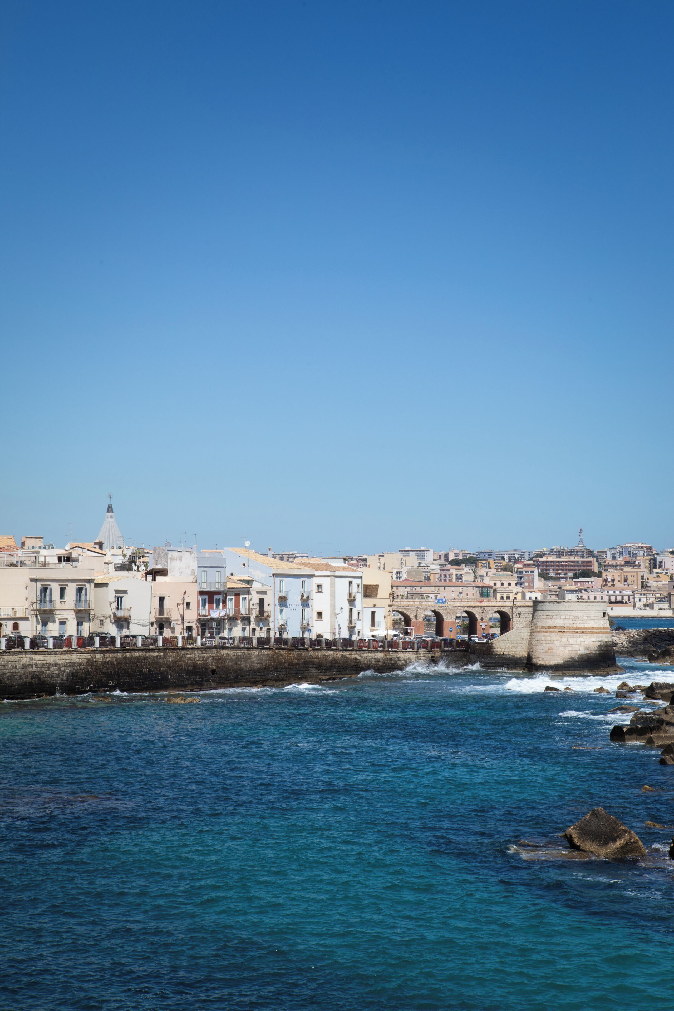 A coastal cityscape with a prominent bridge and buildings along the waterfront.