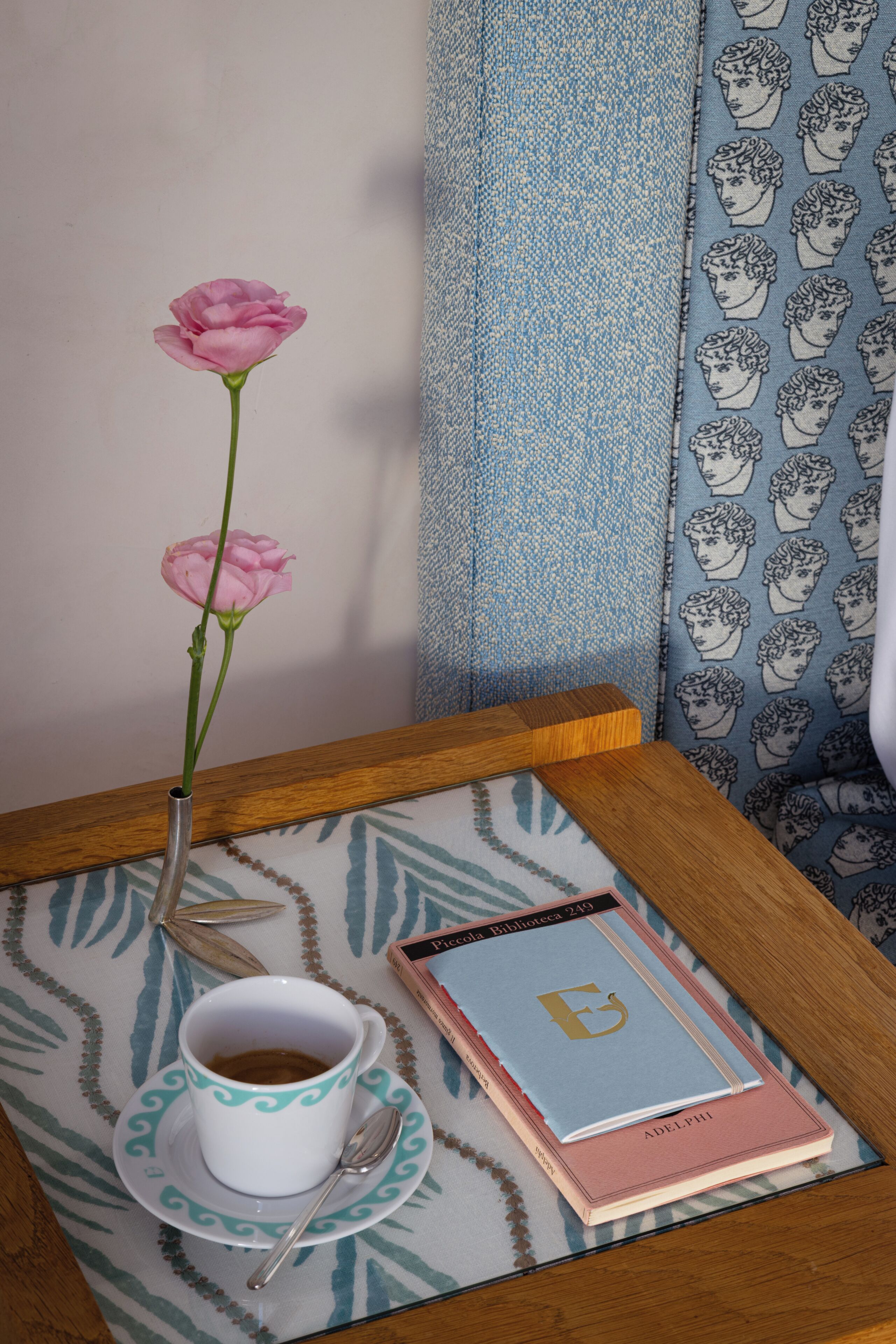 A table with a cup of coffee, a spoon, a book, and a vase with a single pink flower.