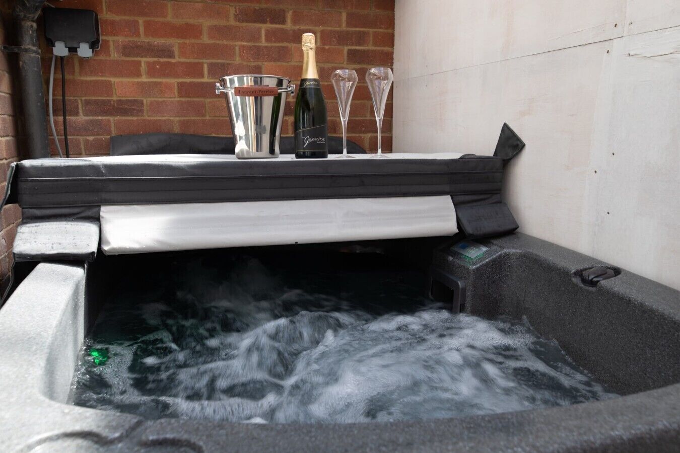 A hot tub with a brick wall and a table with a bottle and glasses.