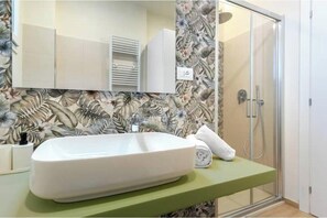 A bathroom with a white sink, a mirror, a shower, and a floral wallpaper. - Hostdomus - Green House (Diano Marina)