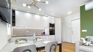 A modern kitchen with white cabinets, a dining table with chairs, and a green accent wall. - Hostdomus - Green House (Diano Marina)