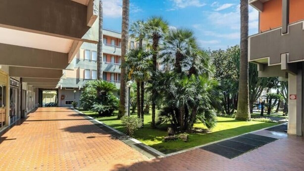 A hotel corridor with a covered walkway, palm trees, and a clear blue sky. - Hostdomus - Green House (Diano Marina)